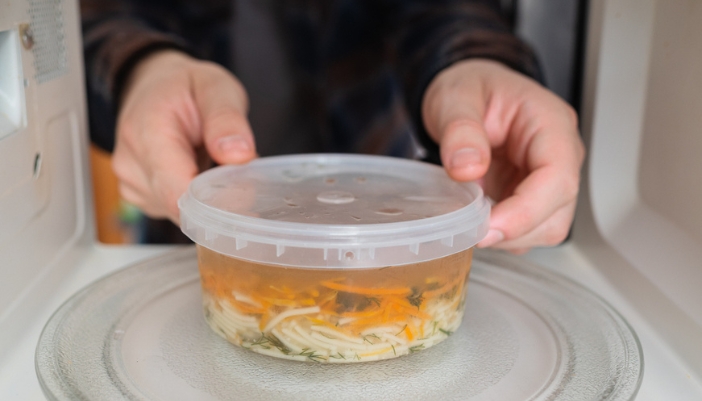 A pair of male hands places a covered bowl of a noodle dish into a microwave.