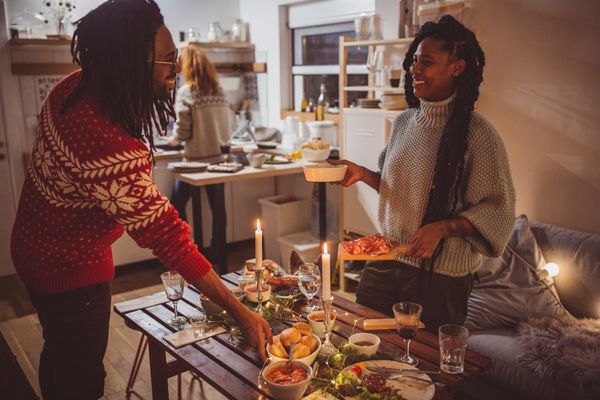 A man and a woman in holidays sweaters smile while setting a table 