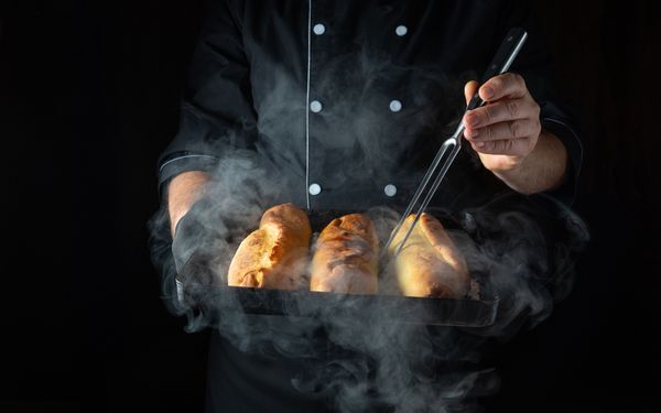 A chef in a black suit holding a tray of steaming baguettes 