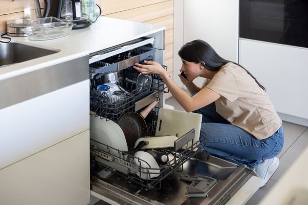 A woman on the phone looking inside of a dishwasher 