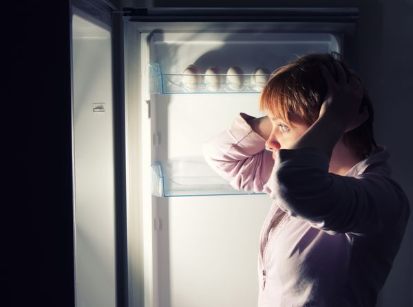 A woman holding hear head looking into a refrigerator 