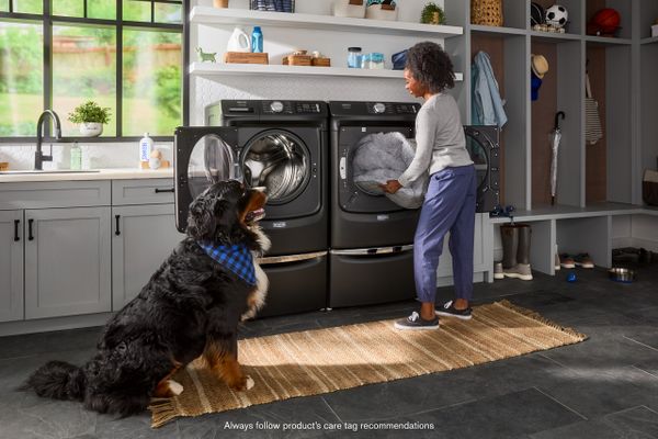 A woman loading clothes into a Maytag Pet Pro washer next to a dog 