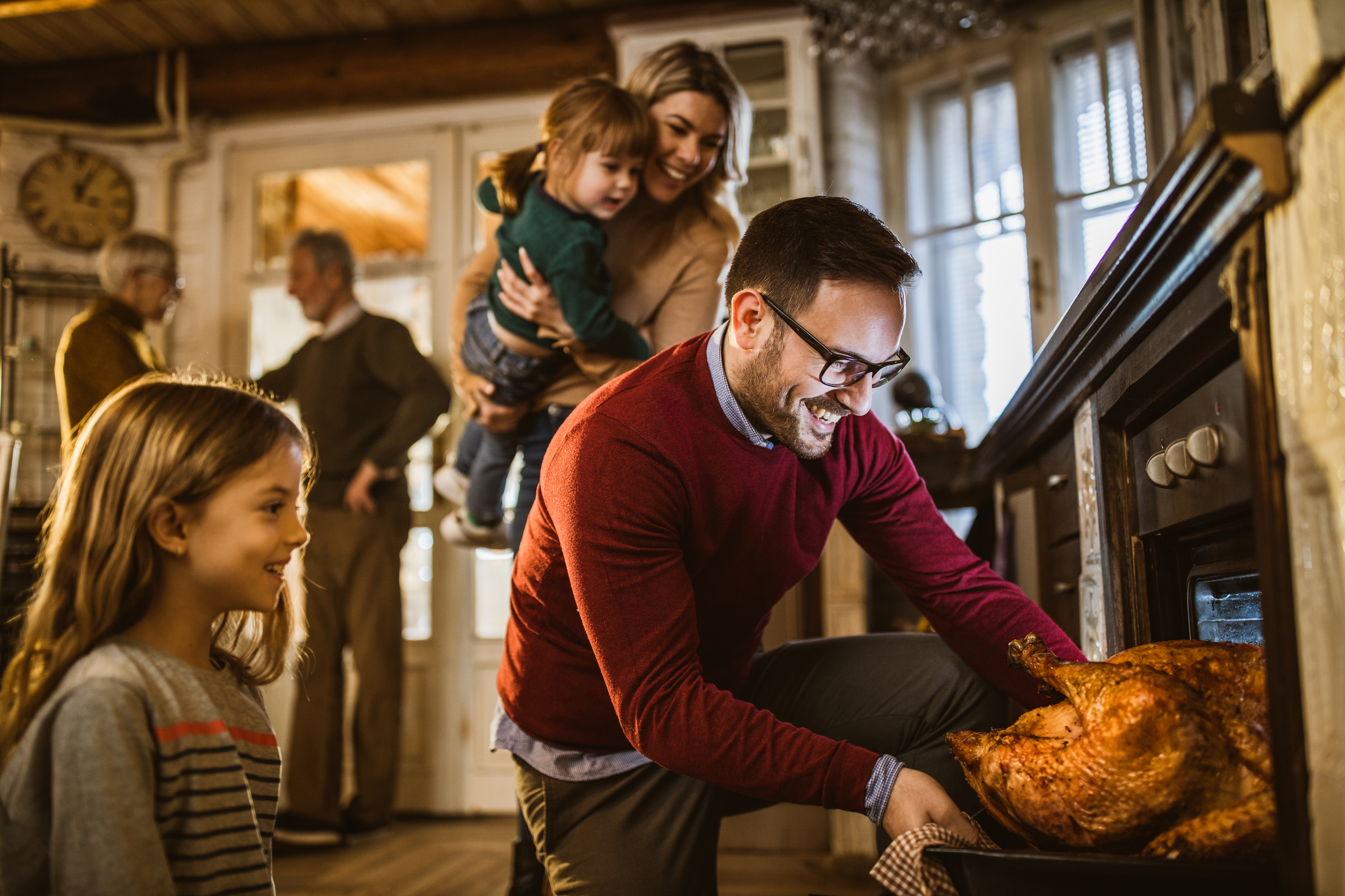 A man pulls out a turkey from the oven while family gathers around 