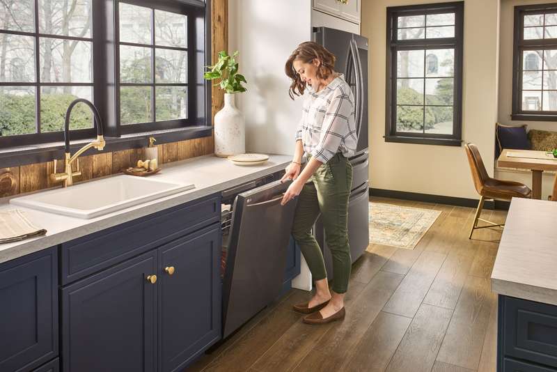 Woman standing in the middle of a sunny, modern kitchen opening up a stainless-steel LG brand dishwasher. 