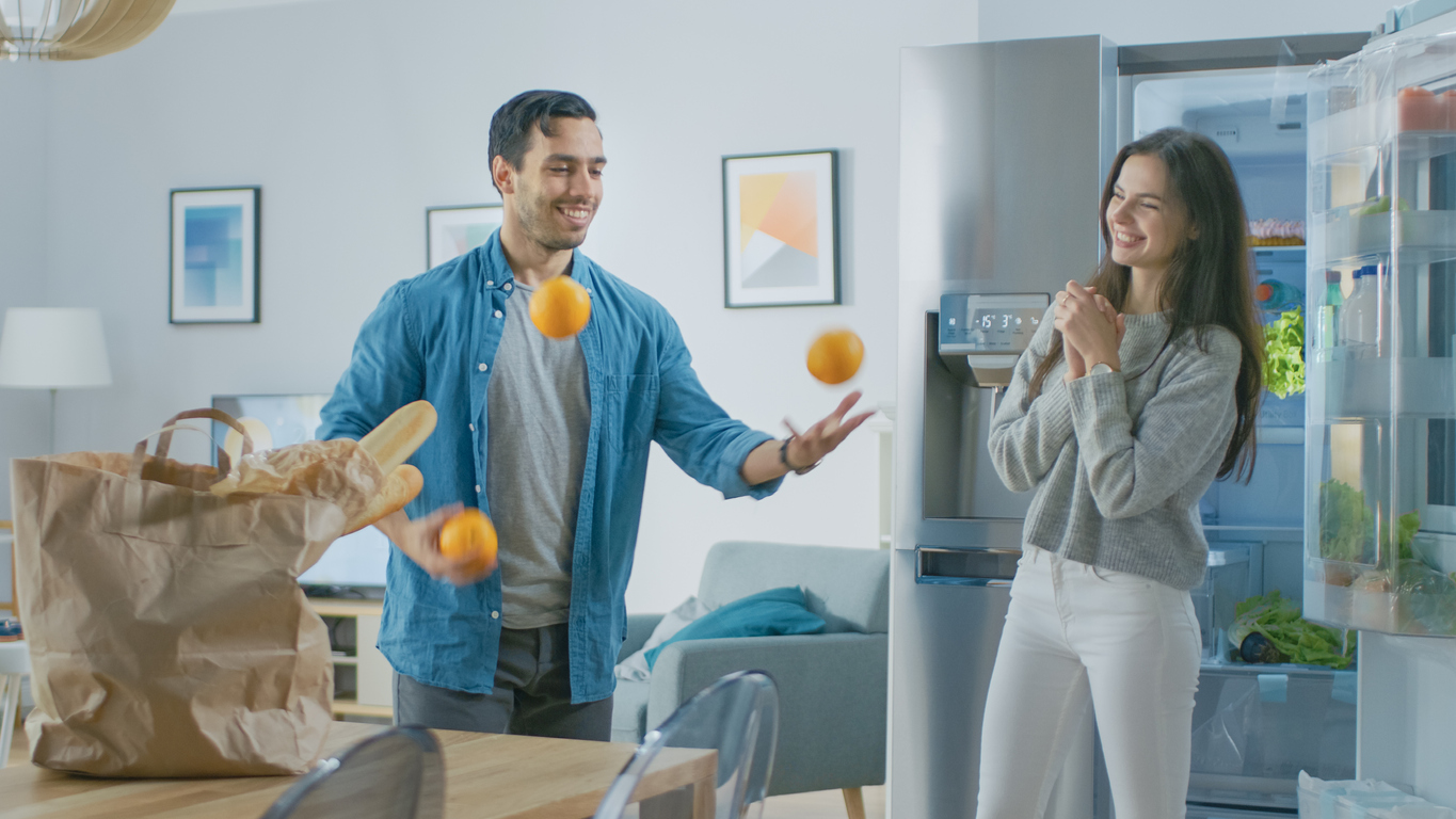 young couple goof around while putting away groceries in refrigerators