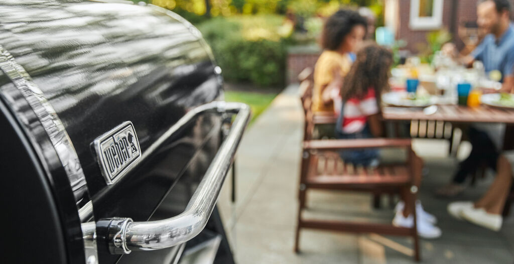 Weber pellet grill shown in the foreground while a family enjoys BBQ in the background 
