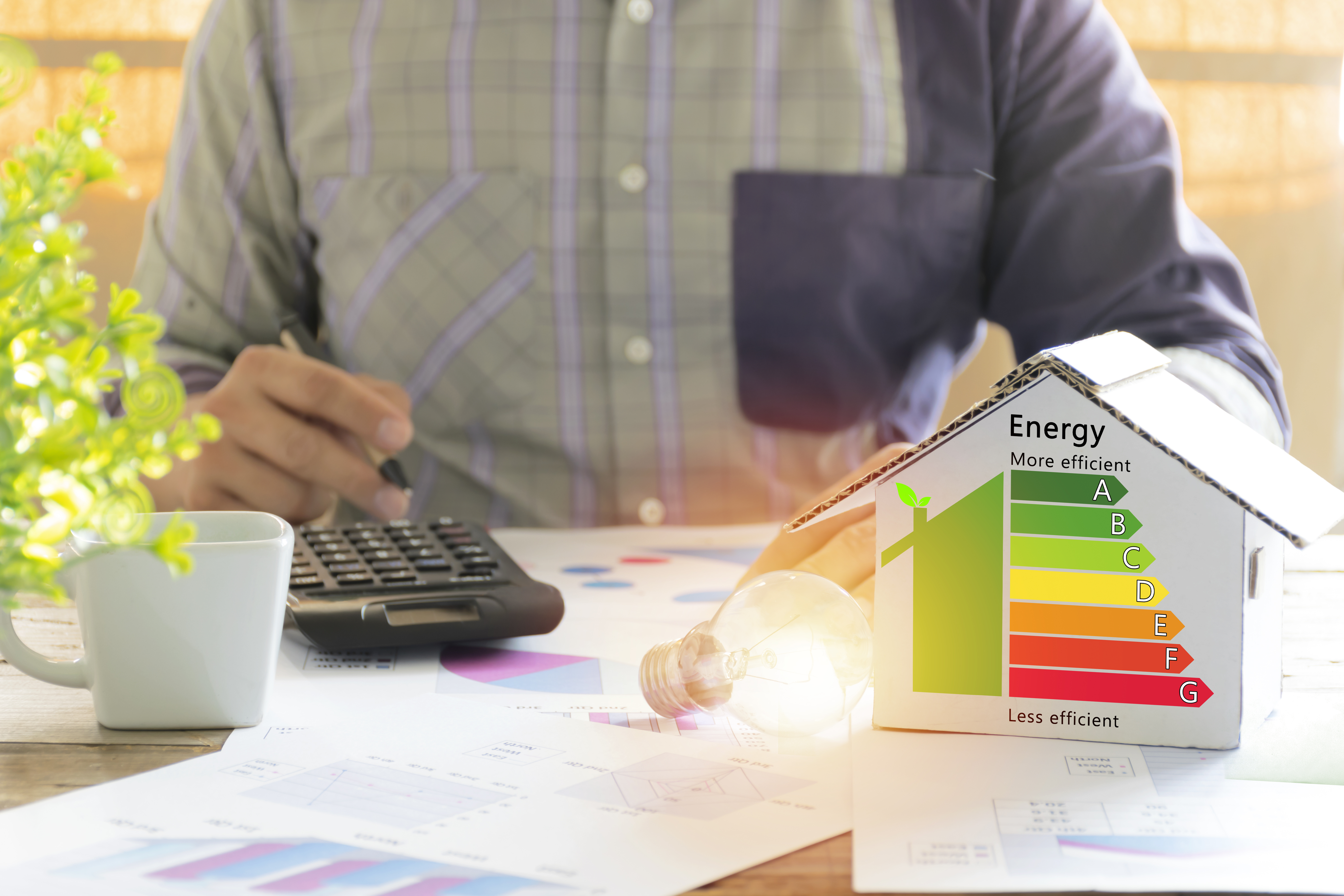 A man using a calculator in the foreground with a cardboard house with a multi-color energy efficiency rating