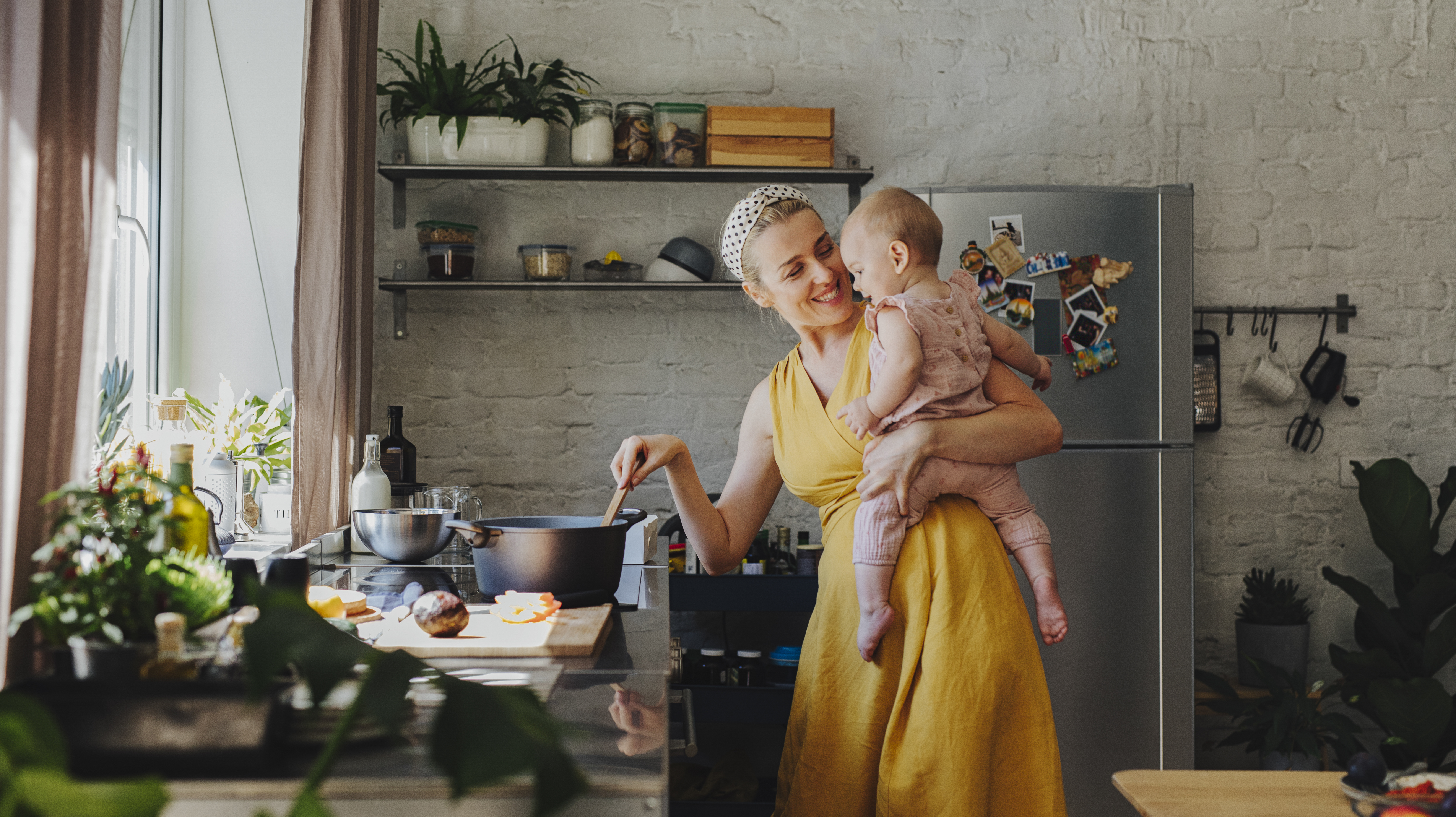 A woman in a yellow dress holding a baby while cooking on a range 
