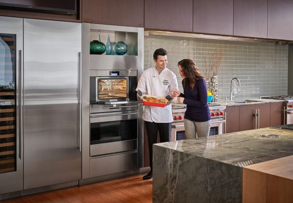 A chef showing a woman an entrée taken from a Wolf oven in a kitchen featuring more Wolf appliances  