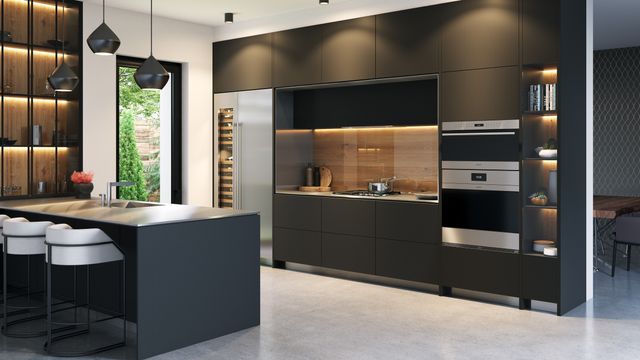 view of a luxury contemporary kitchen with a gray waterfall island in the foreground, and a background of the kitchen with dark gray cabinets, a stainless steel wine cooler and refrigerator, and a double wall oven