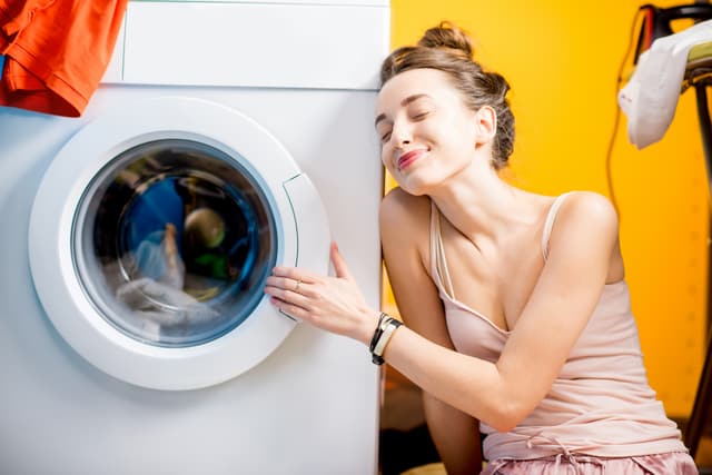 iStock Credit: RossHelen Woman in pink tank top smiles as she hugs front loading washer