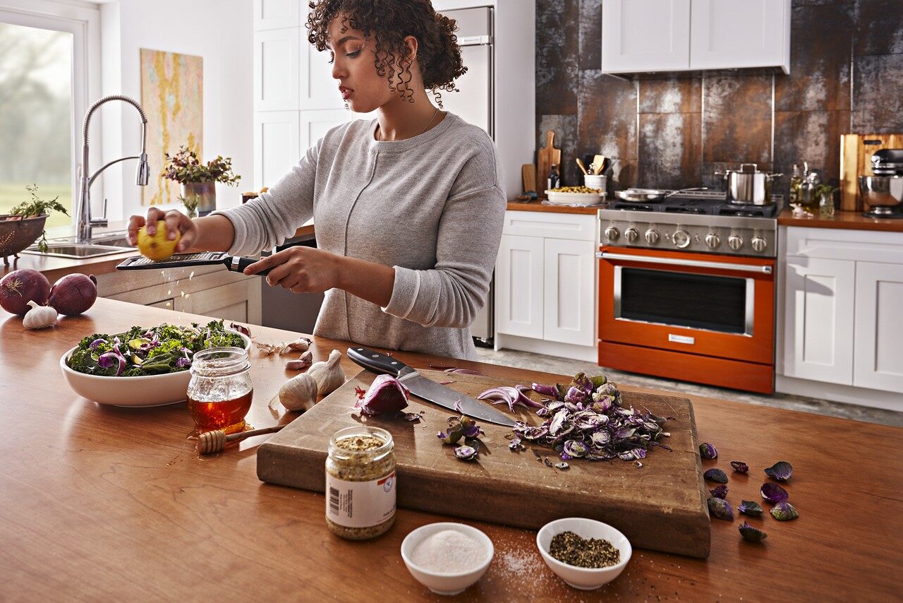 A woman preparing a salad with a shot of an orange KitchenAid range in the background 