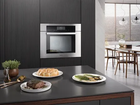 The Miele DGC 7780 CTS wall oven shown in the background of a kitchen with plates of food in the foreground