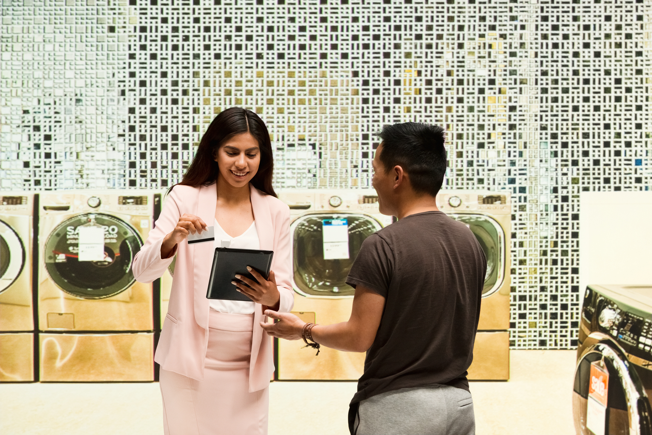 A saleswoman in a pink dress smiling while passing a card through tablet while a man smiles back at her 