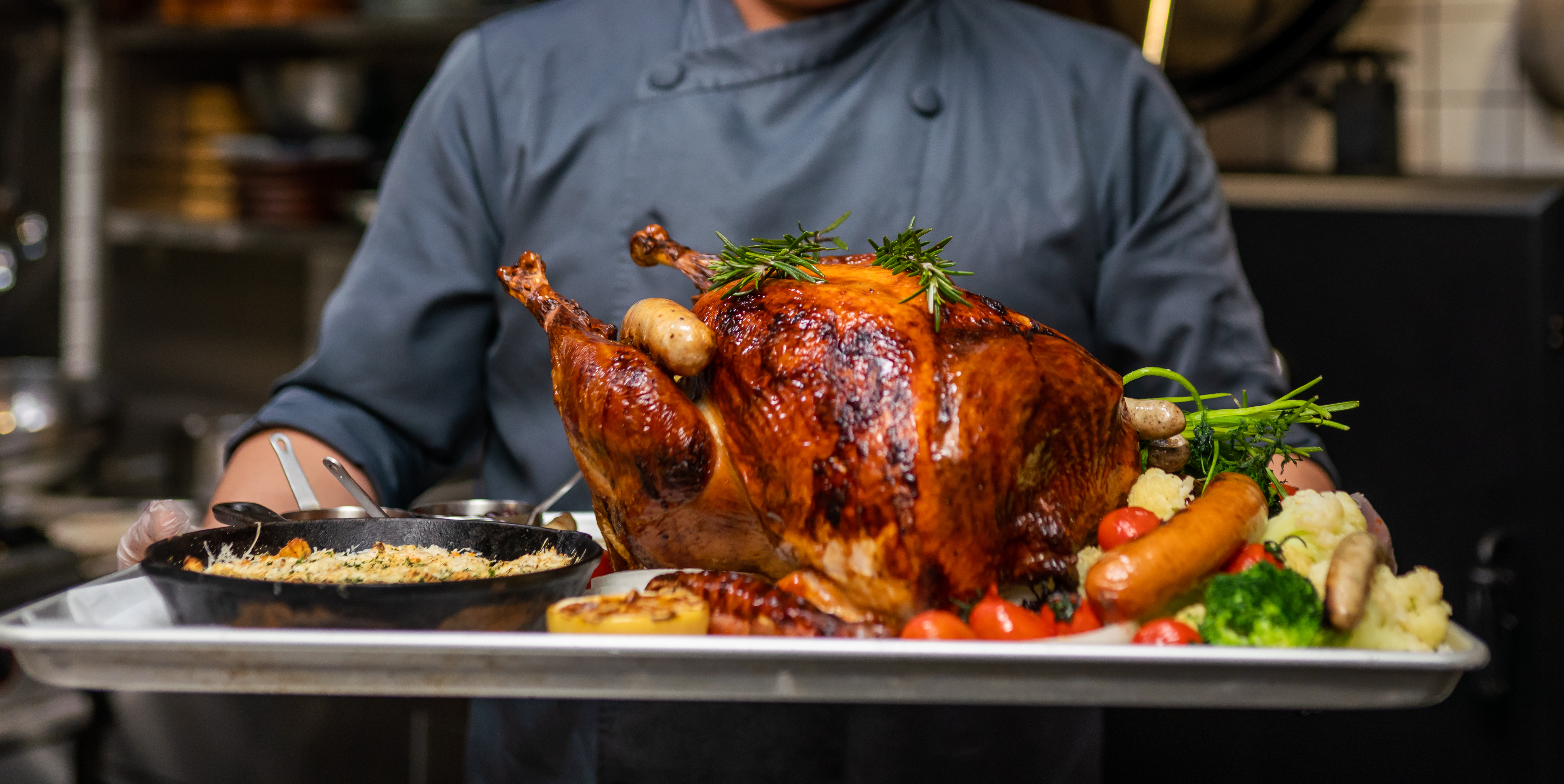 A chef in a blue apron carrying a roasted turkey with sides on a sheet pan 