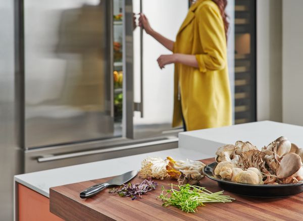 A  woman opening a Sub-Zero refrigerator in the background and a variety of mushrooms in the foreground 