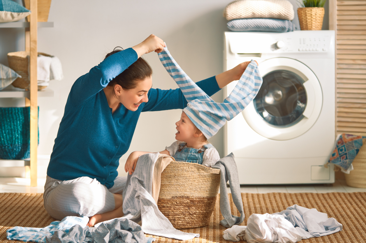 Mother and her child, inside a laundry basket, play with clothes in laundry room