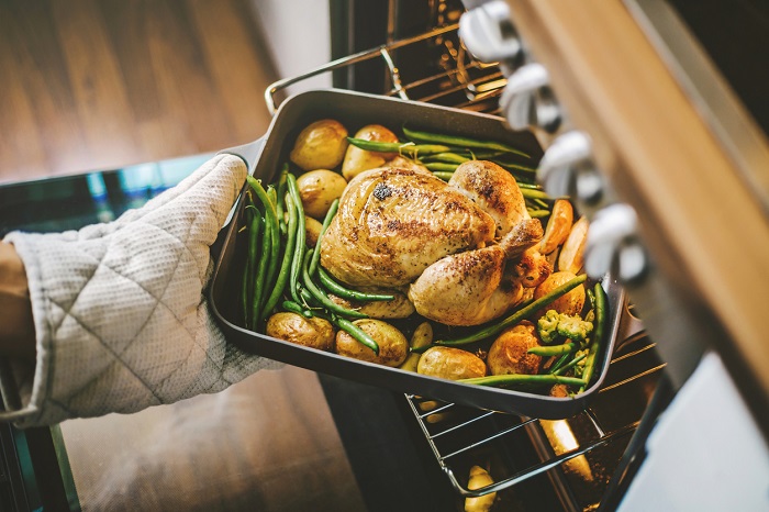 Cook taking ready fried baked chicken with vegetables from the oven.