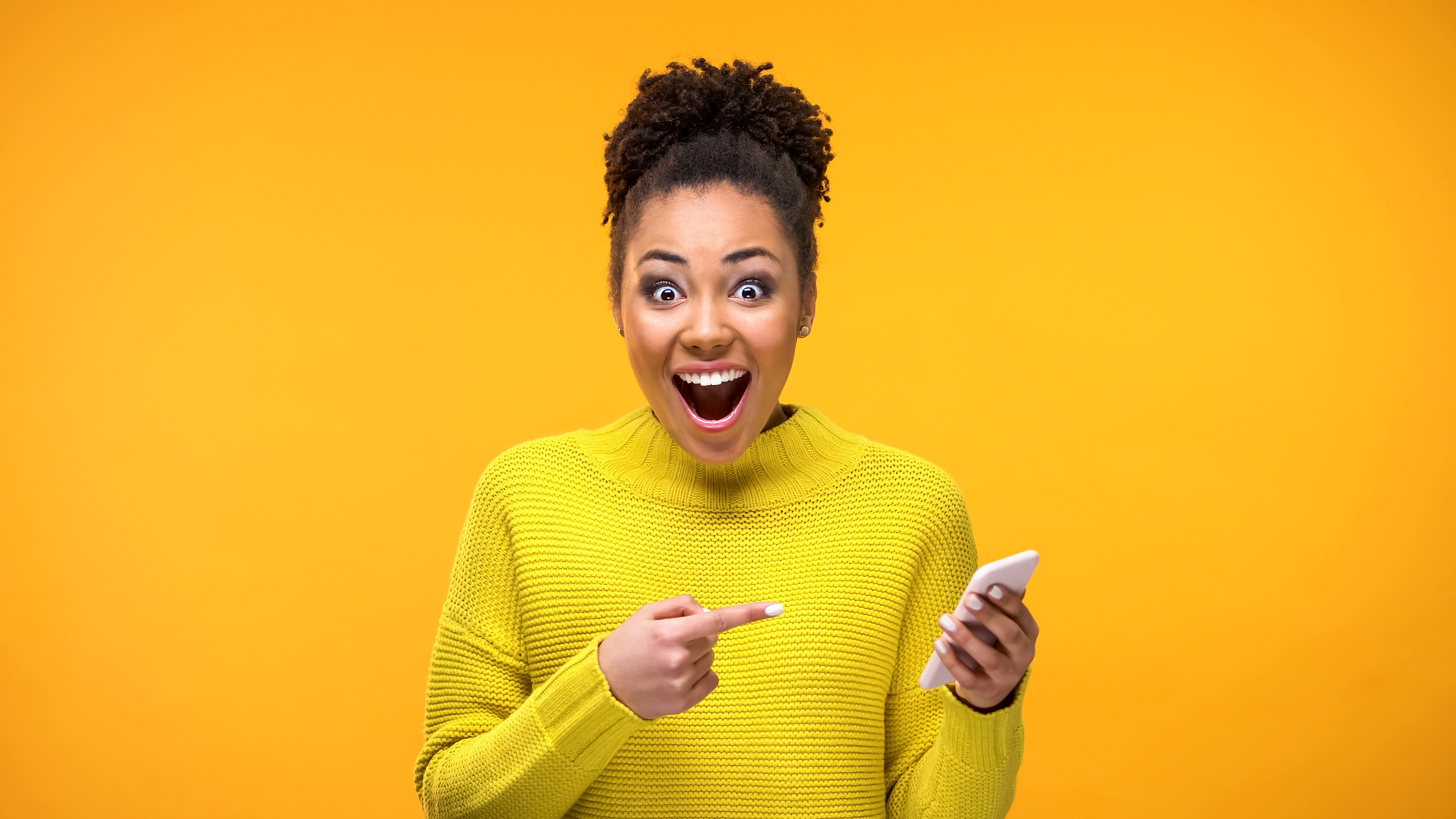 Young woman in yellow sweater smiles and points at smart device while standing in front of mustard-tone background