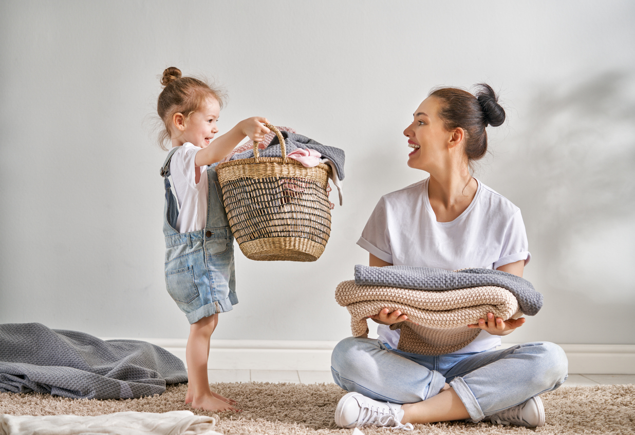 mother sitting cross-legged holding folded sweatshirts smiles at daughter holding laundry basket
