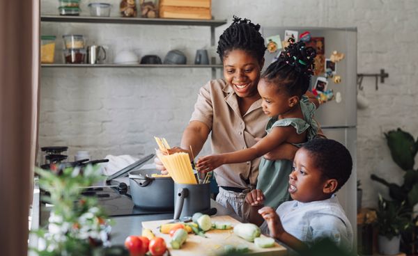 A woman smiling and cooking pasta while holding a child next to another child