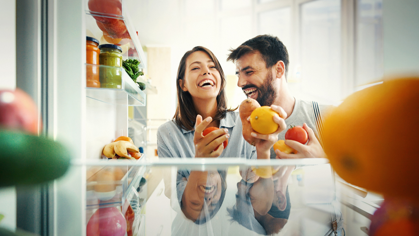 a smiling couple grab fresh fruit from their new refrigerator
