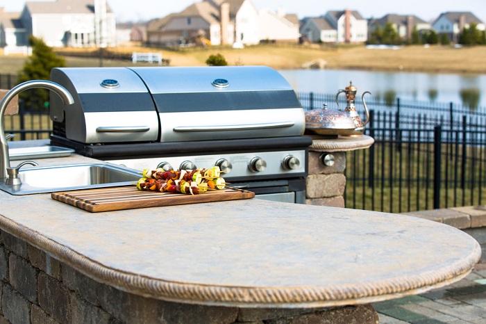 Freshly prepared skewers are waiting on an outdoor kitchen countertop with a stainless steel BBQ grill in the background. 