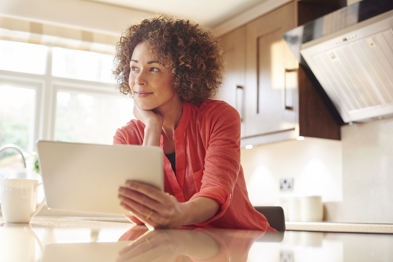 woman in coral shirt holding tablet daydreams in kitche