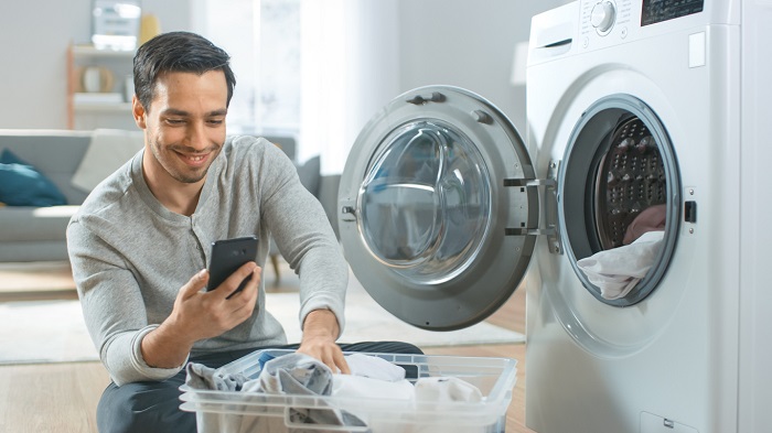 Young man sits in front of a washing machine and is using his smartphone.