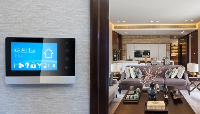 Closeup of smart controls in front of a luxury living room and attached kitchen with aperture lighting