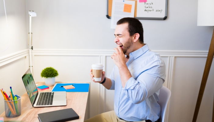 A tired man yawning with a cup of coffee in his hand, trying to work
