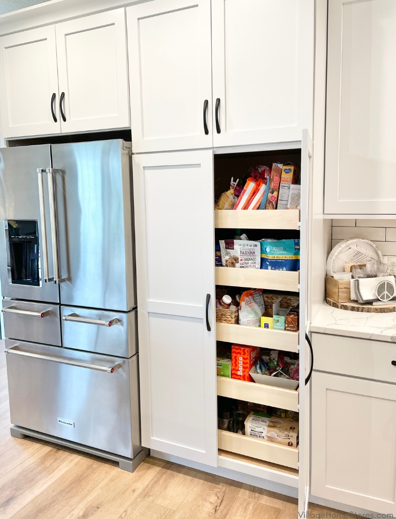 White cabinet kitchen with stainless steel KitchenAid 5 door french door fridge and double door tall pantry cabinet. Right door open on pantry showing sliding shelves stocked with dry goods.