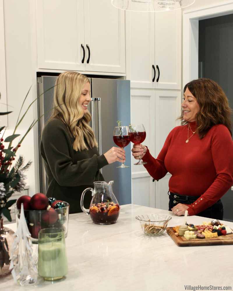 Mother and daughter in white kitchen clinking glasses of red sangria in holiday sweaters from Katsch Boutique