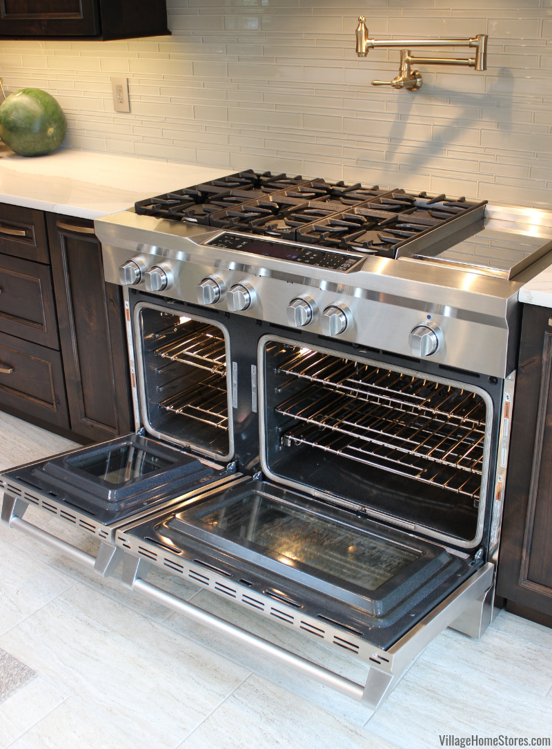 KitchenAid commercial style range with two doors shown open. Installed in dark stained wood kitchen with glass tile backsplash and pot filler.