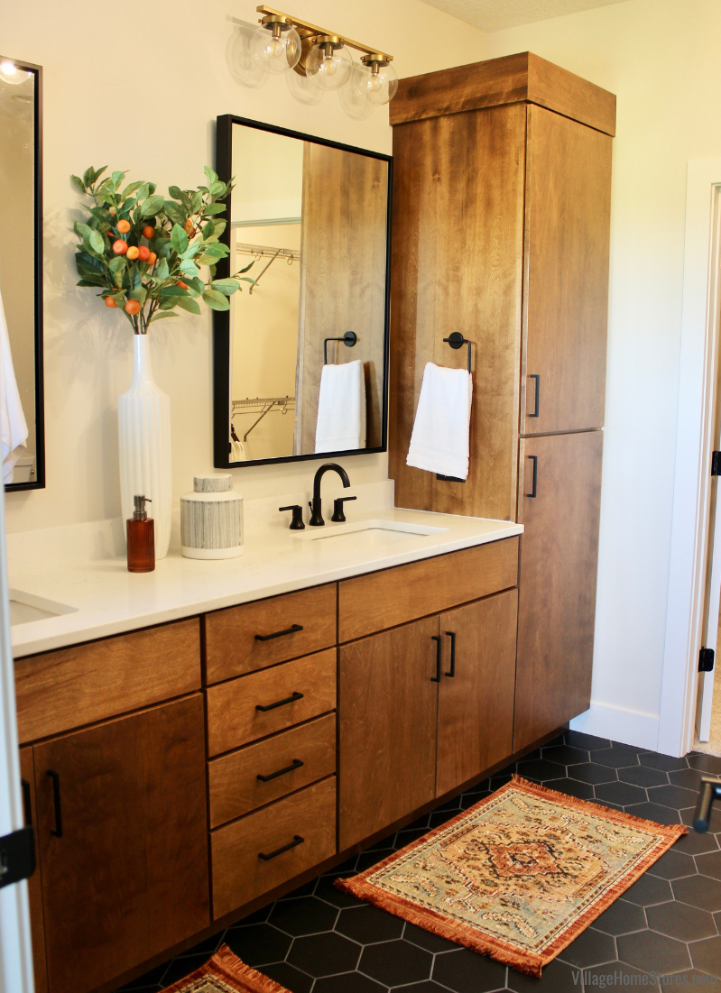 Primary bathroom with medium stained birch wood vanity and linen cabinet. Brushed gold vanity lighting, black framed mirror, and black hex tile floors also featured. 
