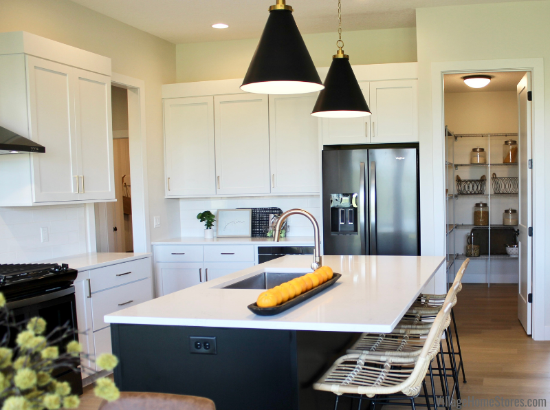 Image of kitchen island with undermounted sink and white quartz countertop. Black stainless refrigerator and walk in pantry in background. 