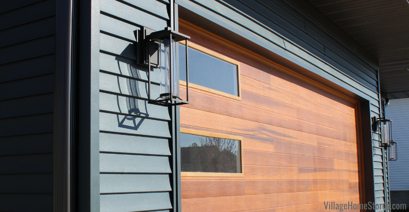 Minka Trescott 17” outdoor lantern light fixture installed on dark gray siding by wooden garage door. 