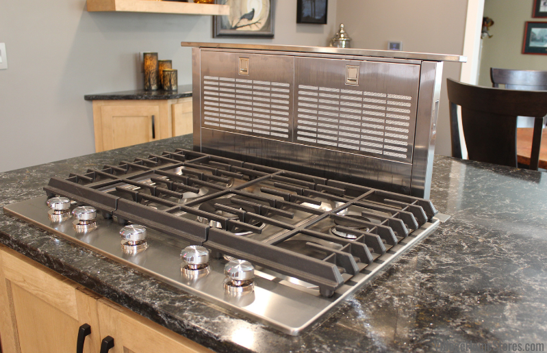 Image of stainless steel gas 6 burner cooktop with downdraft panel raised behind it in kitchen peninsula. Installed in Maple kitchen with dark quartz countertops