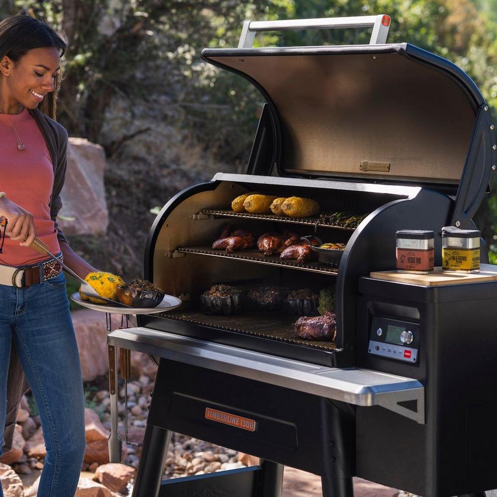 Woman with thongs approaching a Traeger grill with a plate of vegetables.