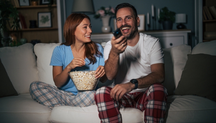 A couple sits on a couch picking out a movie and eating popcorn.