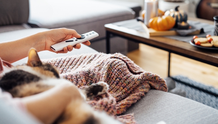 A woman sits under a blanket with her cat and a streaming remote in her hand. 