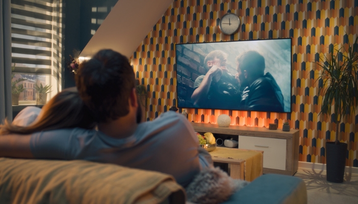 A couple cuddles on their couch at home while watching an action film on TV.