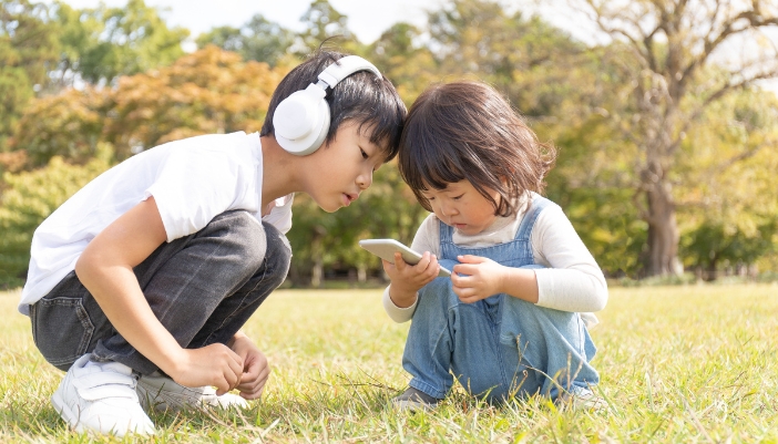 Two kids looking at a smartphone while one kid wears the big headphones
