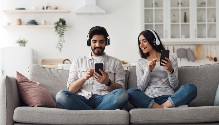 A man and woman sit next to each other on a couch and listen to their headphones.