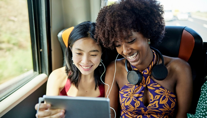 Two women sharing earbuds to watch something on a tablet while one of them has noise-canceling headphones around her neck