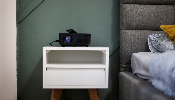 A portable projector sitting on a nightstand next to the bed