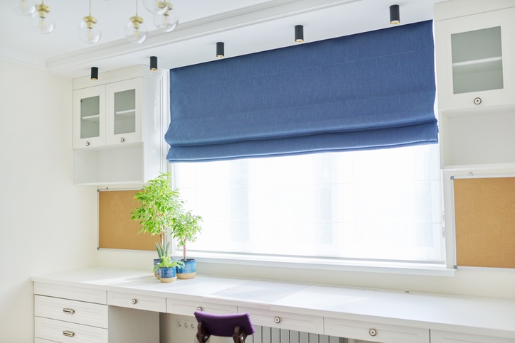 Interior of the room, workplace near window, light furniture, desk, blue roman blind, wall shelves. 