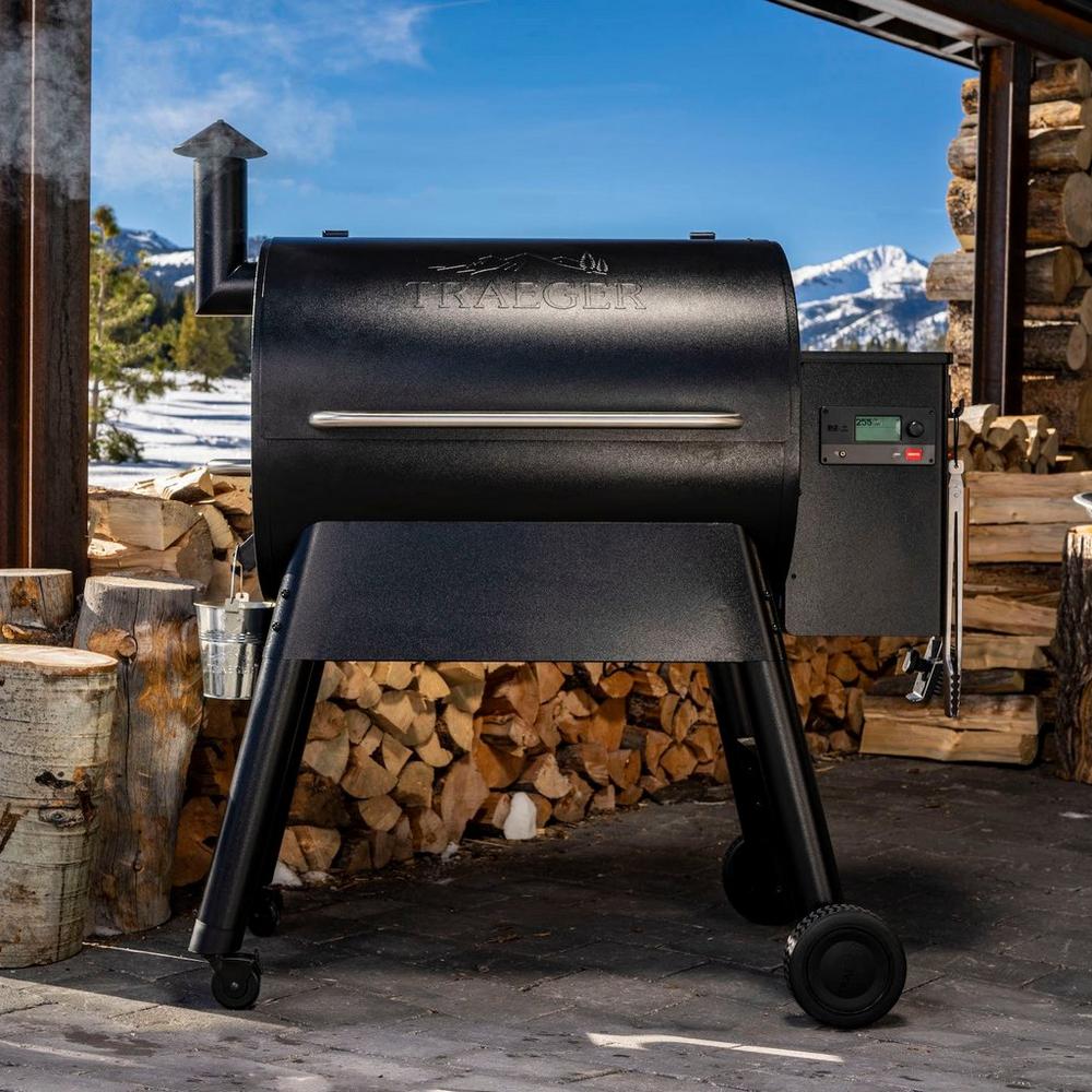 Close up of a Traeger outdoor grill with chopped up firewood and snowy mountains in the background. 