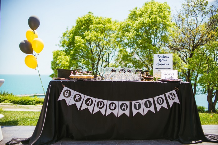 Graduation party table with desserts and glassware, with balloons attached to the side. 