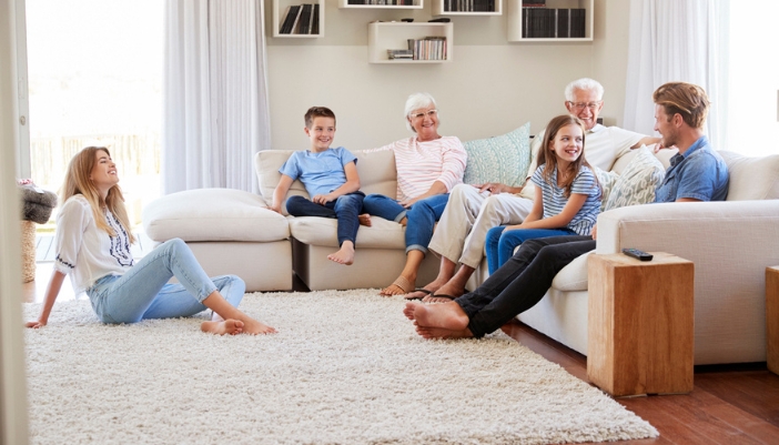 A multi-generational family sits together on a neutral colored sectional sofa.